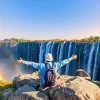 A person with a backpack raising their arms towards Victoria Falls with a rainbow in view.